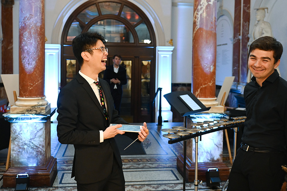 Two students laughing in the RCM’s entrance hall, one holding an iPad. A percussion instrument and another student are behind them.